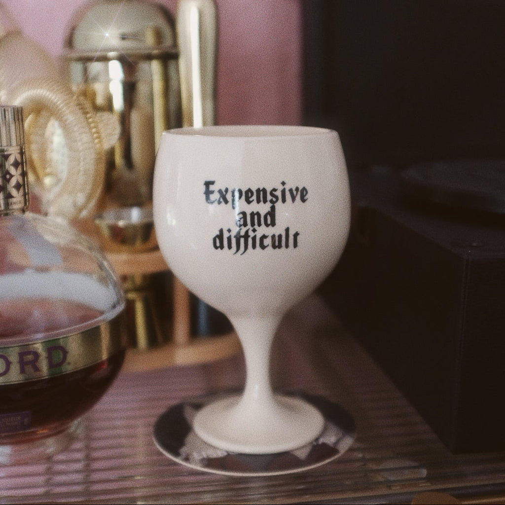 White wine glass with text 'Expensive and difficult' on a bar counter.